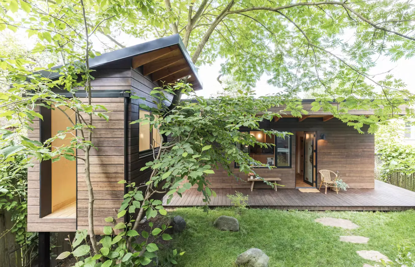 Single-storey backyard ADU with cedar siding and a deck, under a big shade tree in a Winnipeg-style back yard