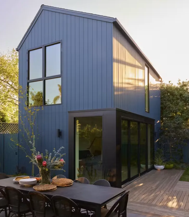 Tall gabled two-storey backyard home in dark blue siding with big windows and a patio
