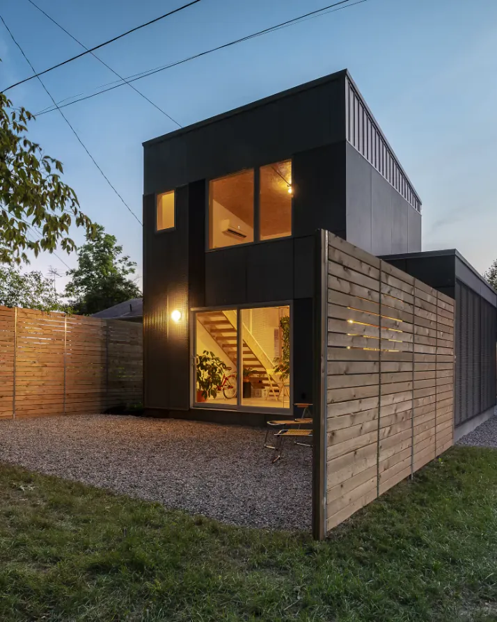 Modern two-storey detached ADU in black siding with lit windows, behind a cedar privacy fence